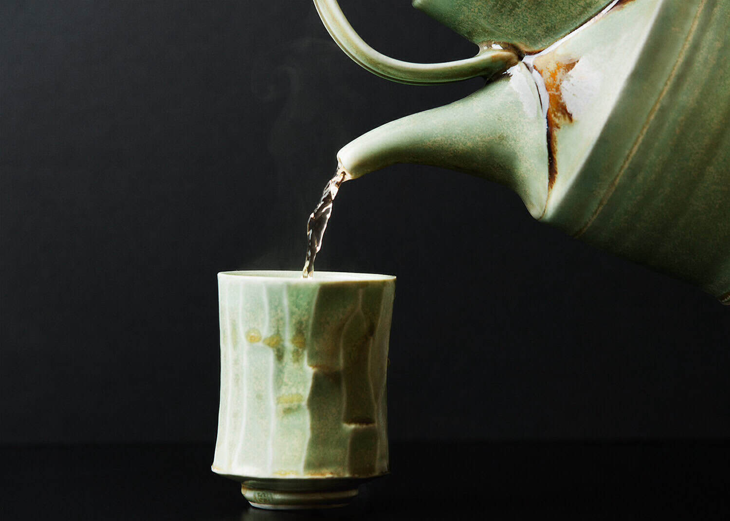Green ceramic tea pot pouring tea into a matching cup.